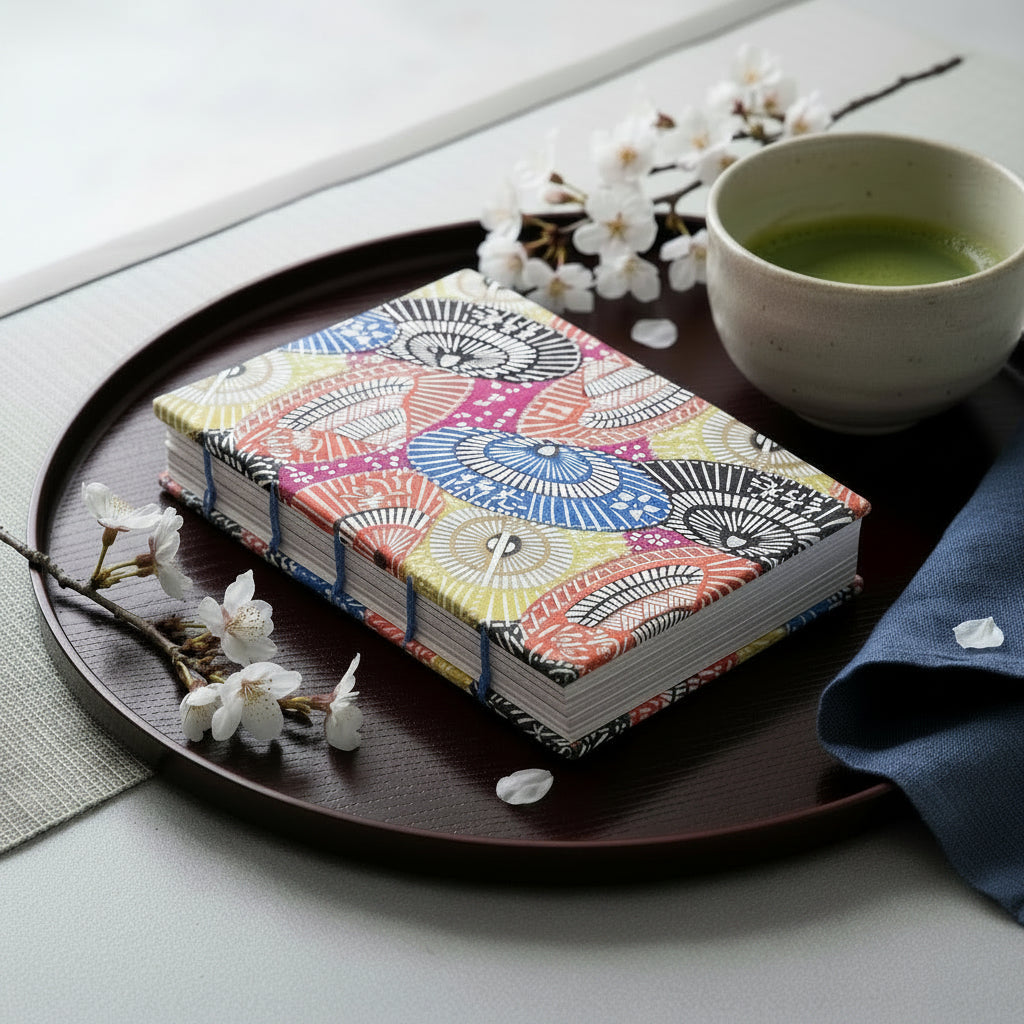 The same handmade book (as the previous image) but placed in a computer-generated setting on a tray, with a cup of matcha, honeysuckle blossoms, and a blue napkin. 
