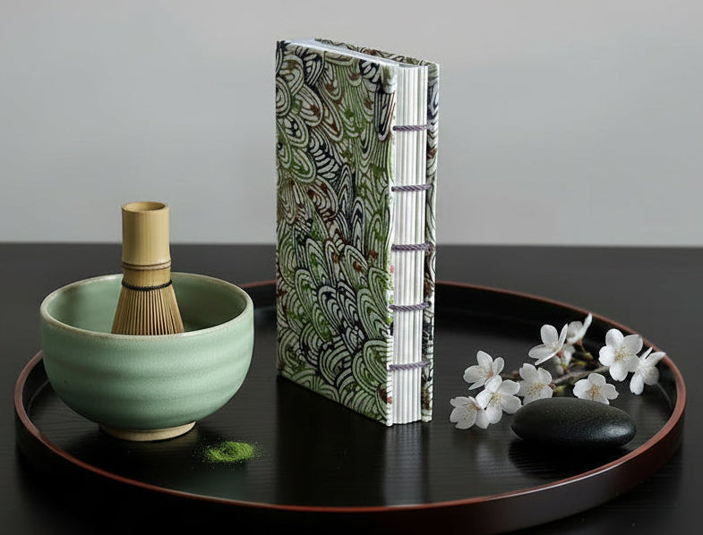 Computer-generated image of a similar book on a tea tray with a matcha bowl, chasen, blossoms, and a smooth black rock.