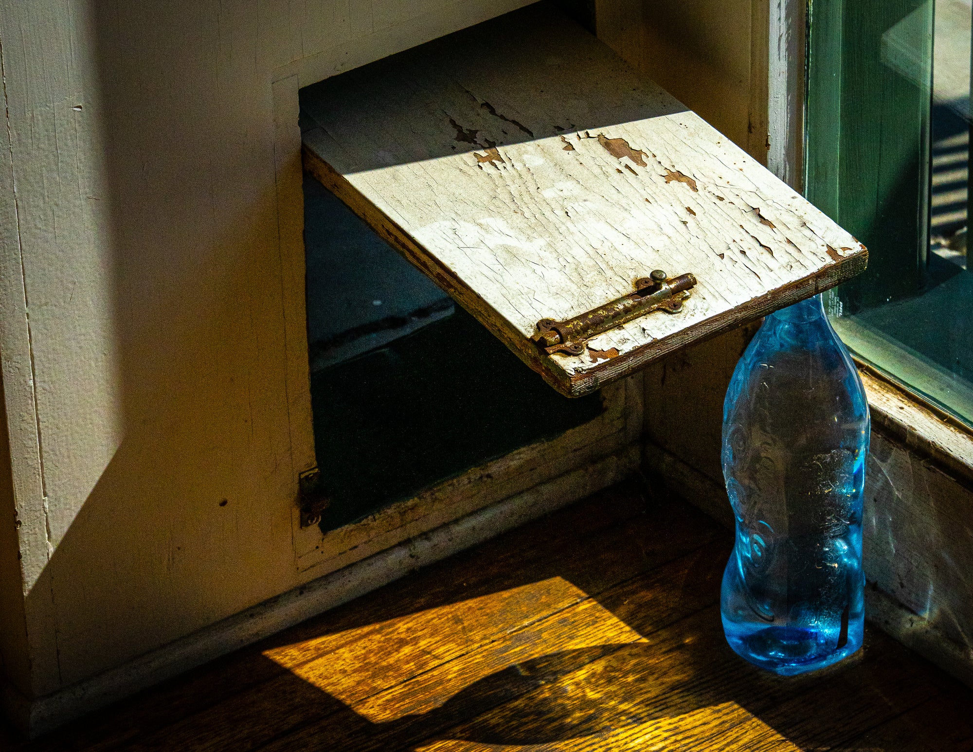 Blue bottle on a wooden floor propping a weathered dog door open, with light from an adjacent window casting shadows; photographer Rick Graves
