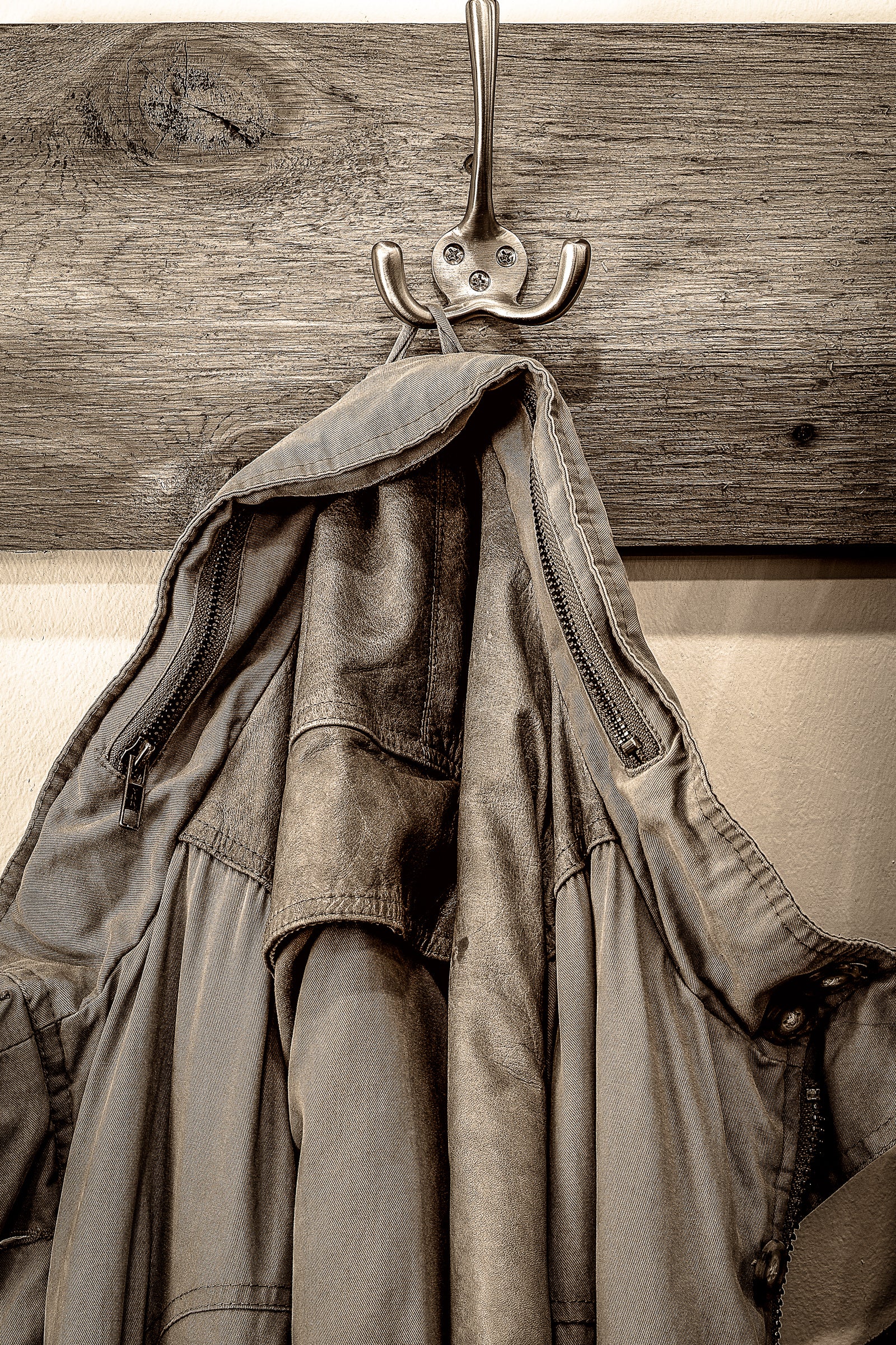 Sepia image of an old canvas and leather coat hanging on a hook mounted in a wooden board; photographer Rick Graves