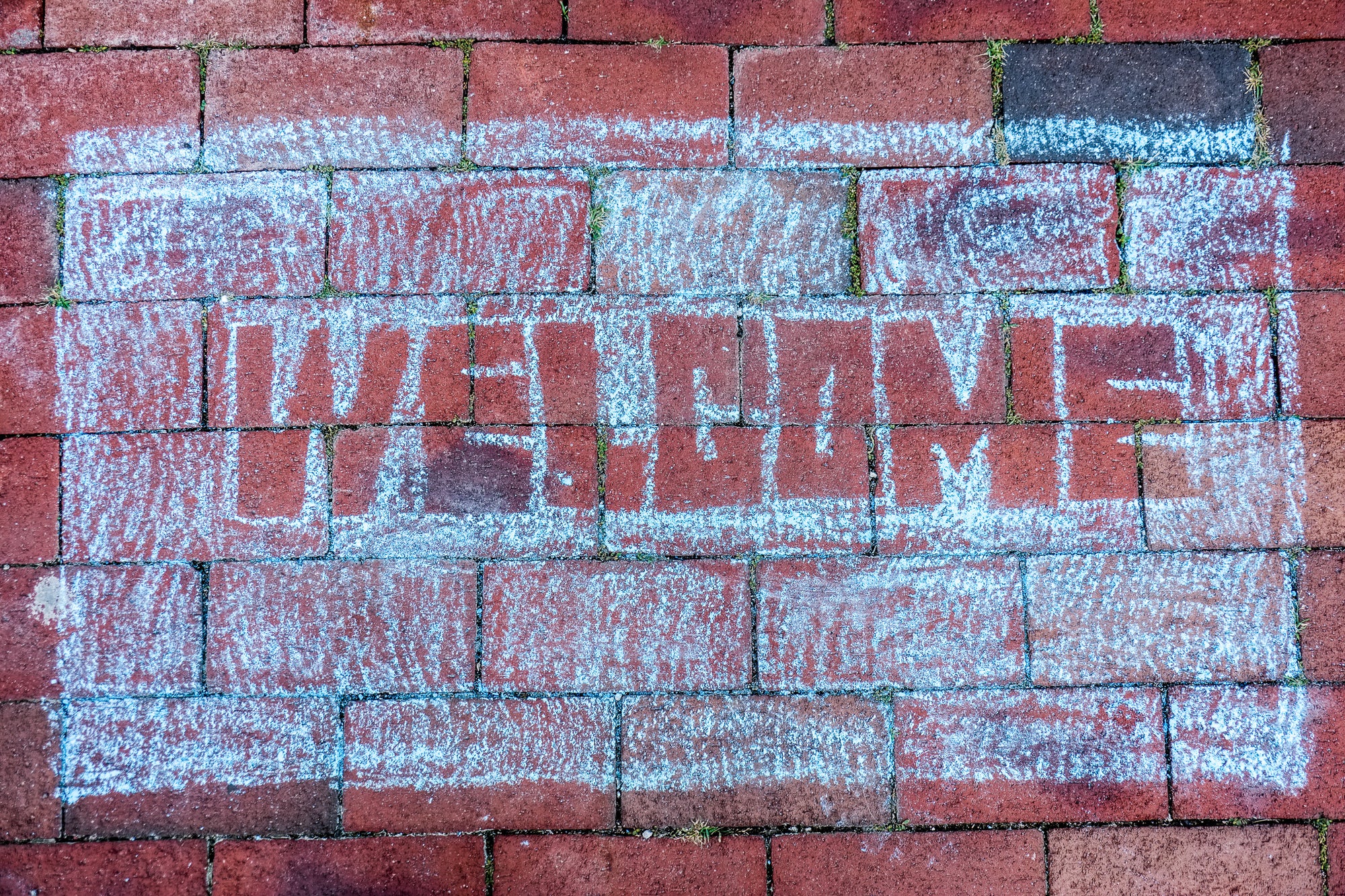 'WELCOME' mat drawn with blue chalk on red brick pavers; photographer Rick Graves