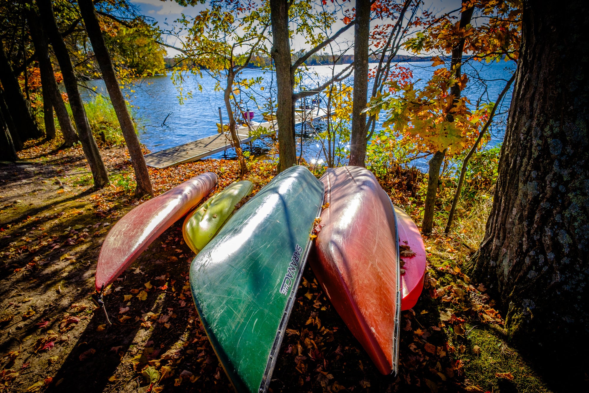 Colorful canoes and kayaks upside-down on shore by a lakeside with autumn foliage. Photographer Rick Graves