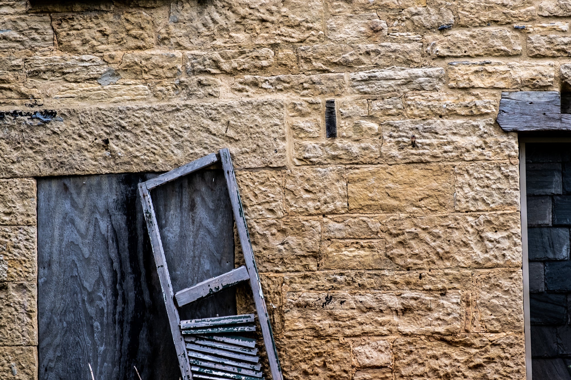 A single weathered wood shutter, on a window covered with old plywood, in a limestone wall; photographer Rick Graves