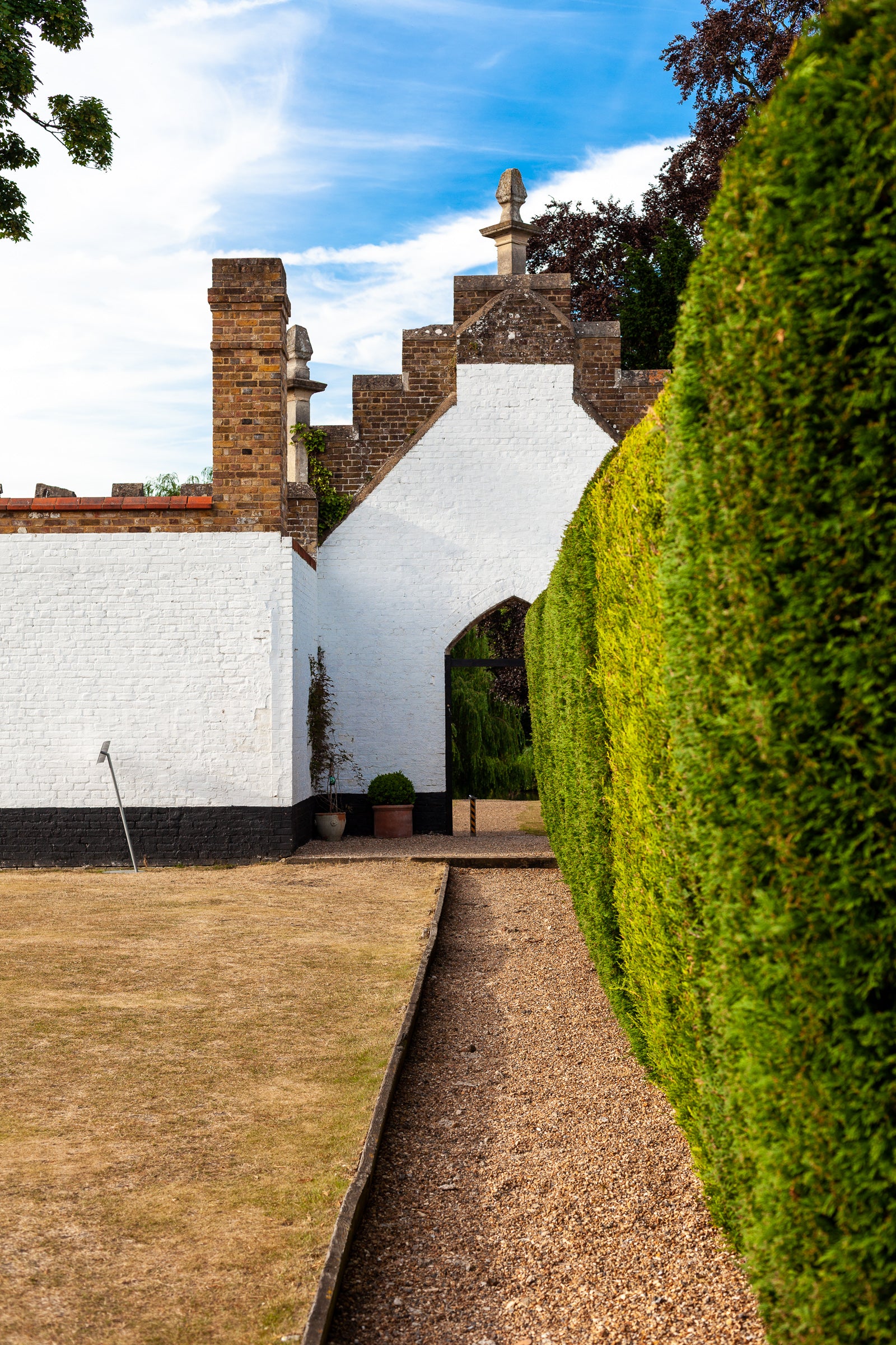 Hedgerow and path leading to a white archway and brick building in the background; photographer Rick Graves