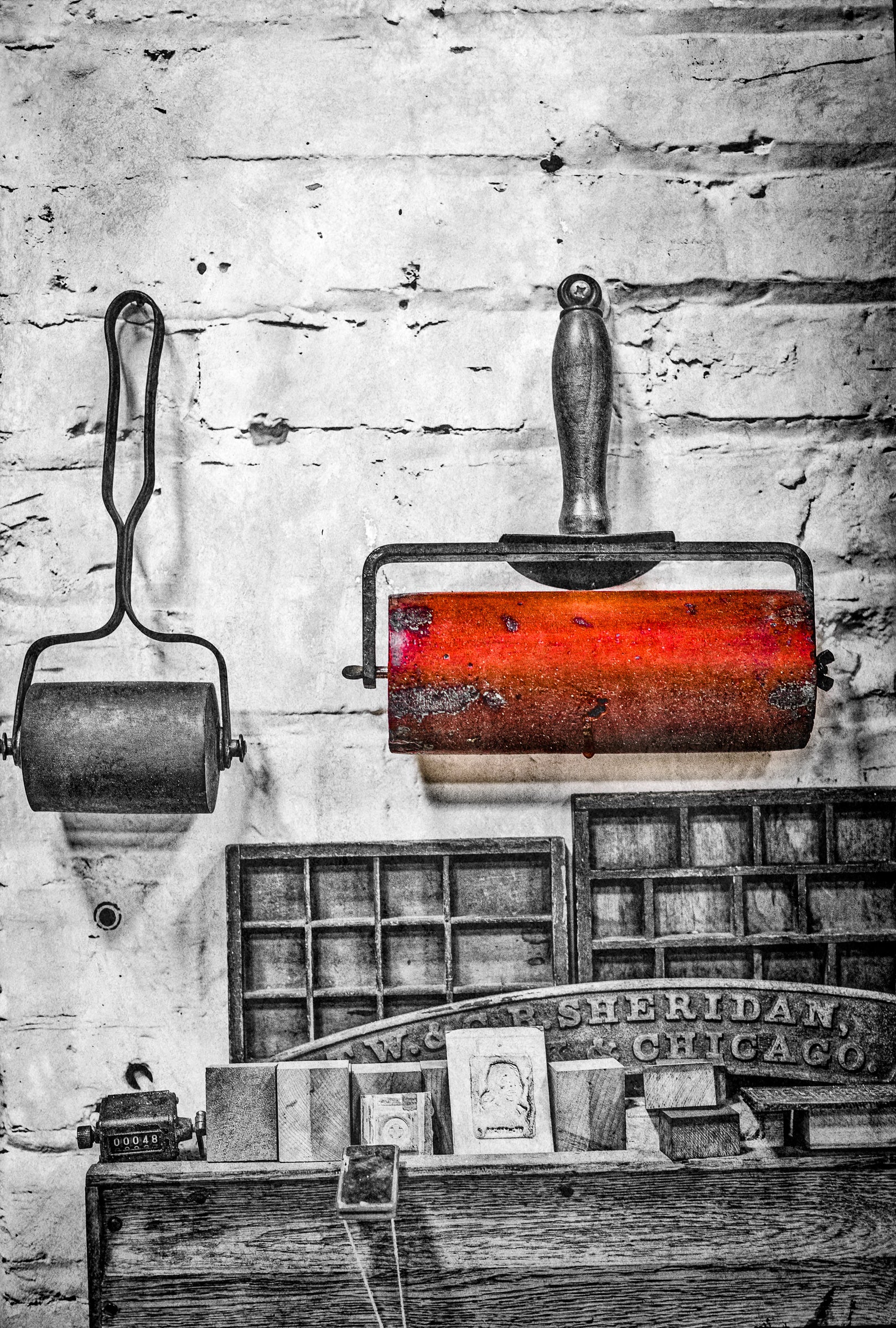 Vintage printing equipment including a roller and type case against a brick wall. BxW with one brayer in red. Photographer Rick Graves