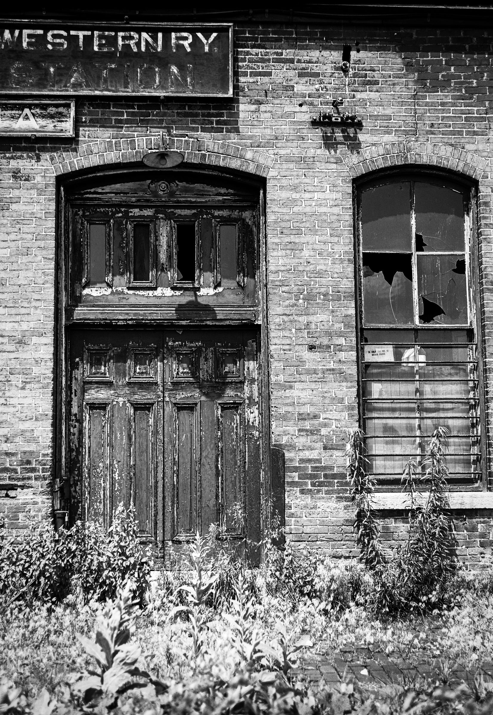 Abandoned building with a wooden door and broken windows, overgrown with vegetation; photographer Rick Graves
