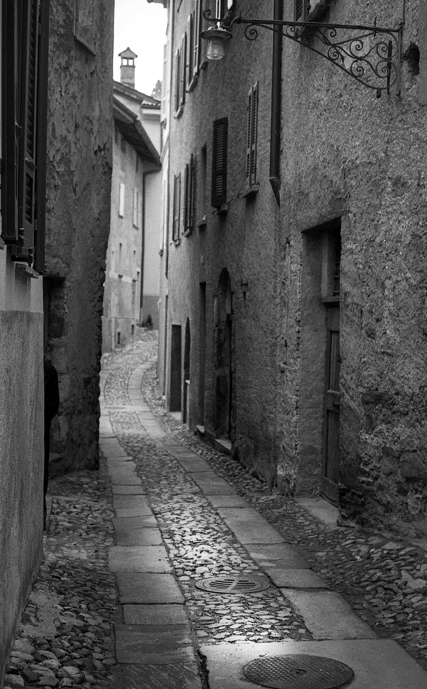 Narrow stone alleyway between old buildings in a black and white photo taken in Rovio, Switzerland; photographer Rick Graves.